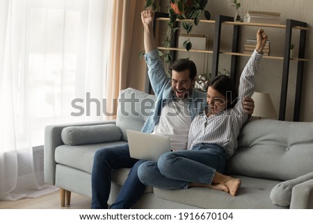 Shocked young family couple cuddle on sofa raise hands scream wow read email with great news on laptop screen. Lucky husband wife celebrate winning lottery getting great offer proposal for purchase