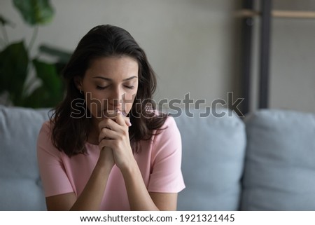 Worried anxious young arabian woman sit on couch praying god with folded hands having life crisis. Nervous millennial mixed race lady trying to concentrate mentally asking supreme forces for help luck