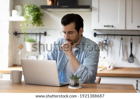 Pensive young Caucasian man sit at table at home kitchen work online on laptop pondering thinking. Thoughtful millennial male look at computer screen browsing wireless internet on modern gadget.