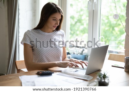 Millennial Caucasian woman sit at desk at home look at laptop screen manage household budget online. Happy young female work on computer calculate pay household expenses expenditures on web.