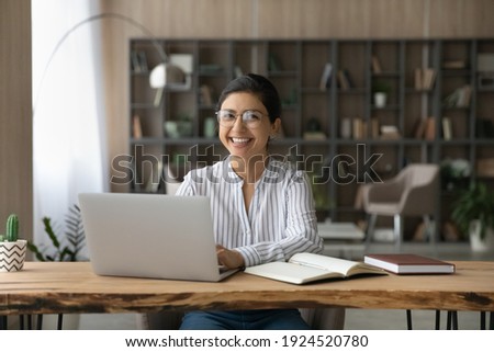 Portrait of smiling millennial Indian female student sit at desk at home study online on laptop. Happy young mixed race woman use computer take distant course or training. Education concept.