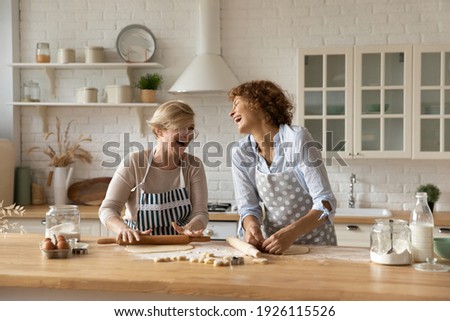 Warm relations. Happy old woman mother pensioner young female daughter grown up kid engaged in baking cookies roll dough at kitchen together laugh have fun. Elderly lady enjoy cooking with adult child