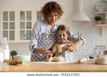 Friendly young mother little daughter cook dessert bakery at modern kitchen knead dough with hands enjoy preparing cookies bread spend time together. Small girl learn to bake with help of caring mom