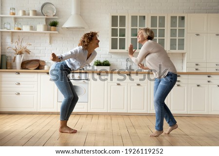 Happy adult daughter and senior mother dancing at modern kitchen after changing interior design moving to new house. Excited mature mom and grown female child celebrate renovation buying new furniture