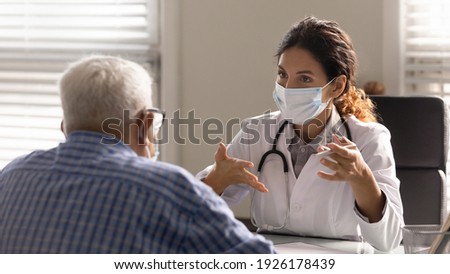 Close up female therapist wearing face mask consulting mature patient at meeting in hospital office, doctor talking, explaining, discussing medical checkup result or symptoms with older man