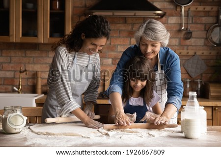 Smiling three generations of Hispanic women have fun baking together with dough at home kitchen. Happy little Latino girl child with young mom and senior grandmother cook pastries or cookies.