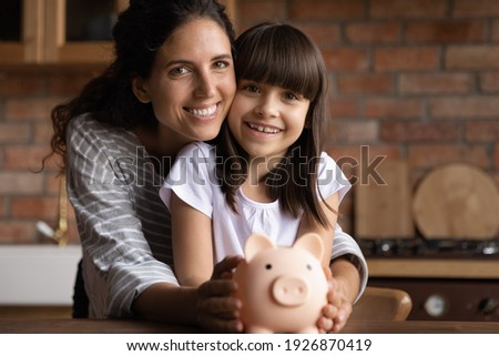 Portrait of happy Latino young mom and small 8s daughter recommend smart living and saving. Smiling Hispanic mother and little teen girl child hold piggybank feel economical making investment.