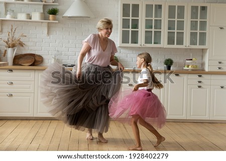 Merry leisure time. Happy energetic grandmother teach ball dance active little child. Caring grandma junior girl grandkid engaged in dancing in funny large fluffy skirts barefoot on warm kitchen floor