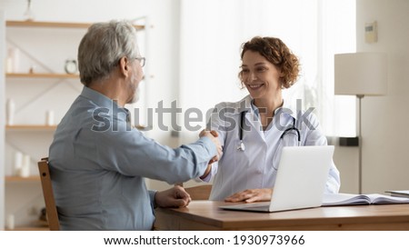 Smiling young female doctor shake hand close health insurance deal with elderly patient at consultation in hospital. Happy woman GP handshake greeting get acquainted with mature man in clinic.