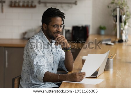 Close up smiling African American man wearing glasses reading good news in letter, holding paper, sitting at table with laptop, received job promotion, money refund, working with correspondence