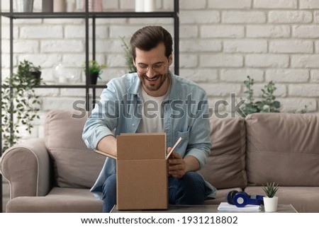 Unpacking parcel. Happy young man postal delivery service client sit on couch at living room open small carton box enjoy goods received. Curious guy online shopper get new order from web store by mail