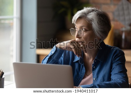 Old age and tech. Thoughtful aged latin female sit at desk work online by laptop from home office. Pensive lady retiree learn to use internet study computer ponder on next step distracted from screen