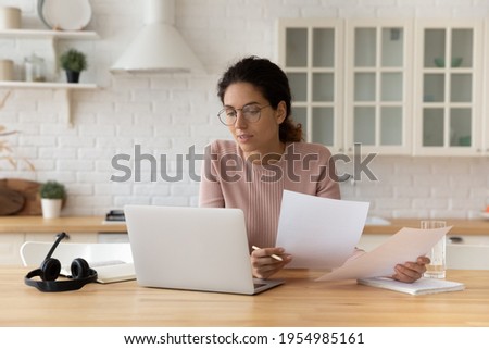Thoughtful hispanic female sit at home workplace hold financial documents look on pc screen check data. Serious young woman think on business offer compare terms conditions in paper letters and online Foto stock © 