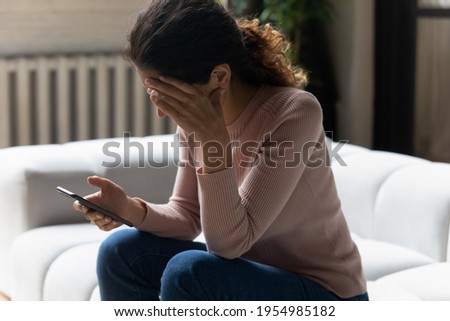 Out of the blue. Frustrated millennial latin woman sit on sofa read sudden bad news from cell screen cover face with palm in desperate gesture. Stressed young lady get nasty blackmail message on email 商業照片 © 