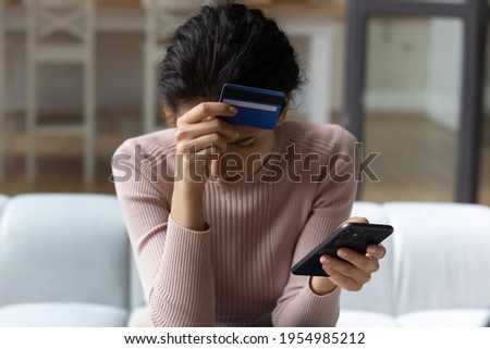 Upset latina female internet shopper sit on couch hold phone with opened web shop ebank page suffer of overspending money from card account. Frustrated young lady lost savings as scam operation result