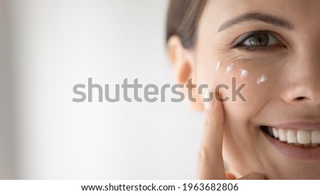 Happy young woman putting moisturizing nourishing cream, cleansing lotion on smiling face, applying cosmetics for keeping healthy fresh skin. Cosmetology, skincare concept, Close up, cropped portrait
