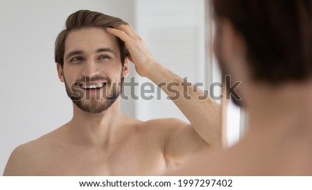 Happy handsome young guy combing smooth brown hair with fingers at mirror in bathroom, looking at reflection with toothy smile. Man satisfied with haircare cosmetic product, enjoying bath routine