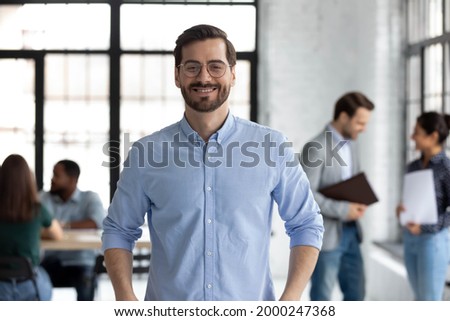 Portrait of happy millennial male business owner in modern office. Businessman wearing glasses, smiling and looking at camera. Busy diverse team working in background. Leadership concept. Head shot.