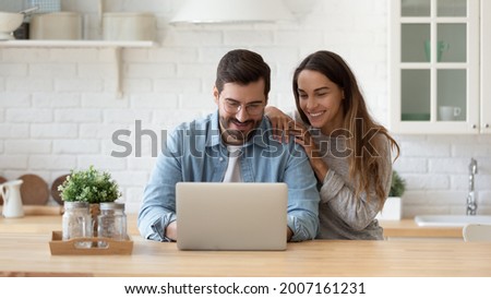Attractive 35s couple sit at table in cozy kitchen looking at laptop screen, buying goods, tickets online, using e-commerce retail e-services feel satisfied, enjoy comfort usage of modern technology