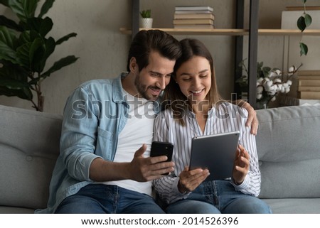 Young 30s couple sit on sofa use diverse gadgets holding smart phone and tablet device. Husband show explains to wife new mobile apps, comparing applications enjoy modern tech usage on weekend at home