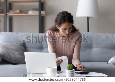 Serious female in glasses paying utility bills, taxes, rental fees use online services e-bank app on laptop. Young woman work from home sit on couch hold paper invoice calculate expenses, make report Foto stock © 