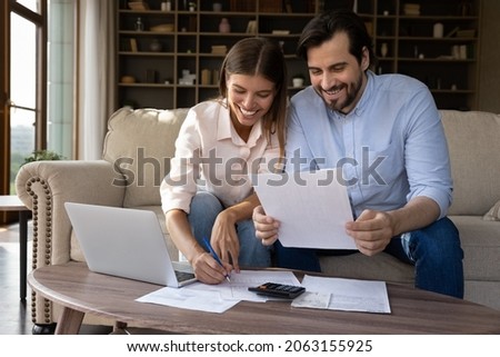 Happy young family couple managing household budget, enjoying planning investment, analyzing paper bills, making payments online in e-banking computer application, doing financial paperwork at home.