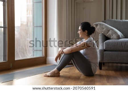 Dreamy Indian ethnicity woman in sports pants and t-shirt looks out window relax after work out training sit near heating grid with ventilation by floor in hardwood flooring. Modern home, rest concept 商業照片 © 