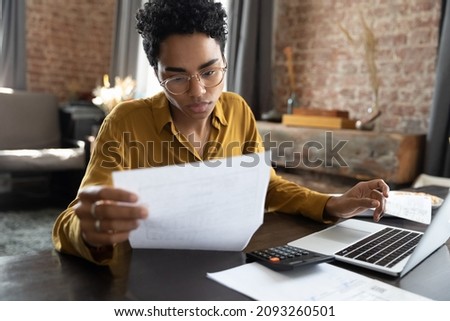 Focused young African American woman in eyeglasses looking through paper documents, managing business affairs, summarizing taxes, planning future investments, accounting alone at home office. Imagine de stoc © 