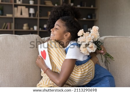 Loving African daughter and young mom hugging holding carnation bouquet and postcard celebrate International Womens Day sit on sofa cuddling feeling attachment showing love. Happy Mother Day concept 商業照片 © 