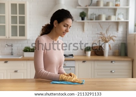 Housewife standing in modern cozy kitchen wear protective glove wipes wood dining table surface with microfiber rag, use professional cleaning products for home perfect tidiness, housekeeping concept Imagine de stoc © 