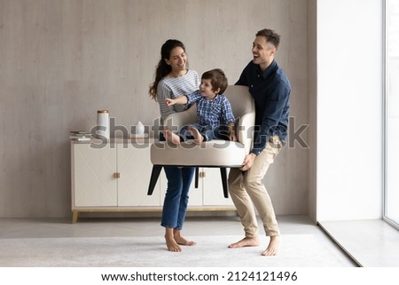 Couple of happy funny parents holding cheerful little son sat in armchair. Family moving into new house, carrying furniture, feeling joy. Mom and dad playing active games with kid. Relocation concept Foto stock © 