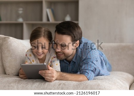 Cheerful millennial father hugging sweet gen Z daughter girl lying on sofa at home, using learning application, tablet computer, playing online videogame on digital gadget, reading ebook on internet Imagine de stoc © 