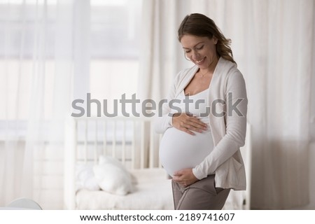Young 30s happy pregnant woman in casual clothes embracing caressing her big belly, standing pose in light nursery at modern cozy home in last stages of pregnancy. Expectant mother, motherhood Stockfoto © 
