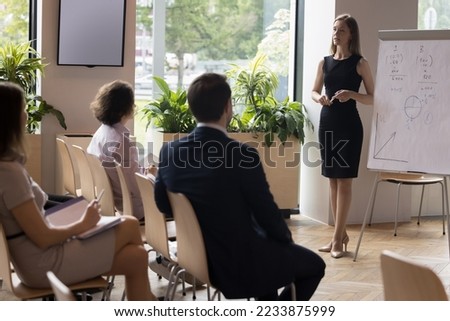 Female business coach make presentation for clients, stand near flip chart with data, sales results and forecast, provide information to staff take part in educational, informative training in office 商業照片 © 