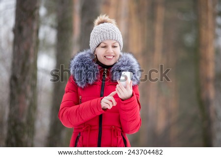 Happy beautiful girl in knitted hat and red winter coat, outdoors, using mobile phone, touches the screen of smart phone, texting