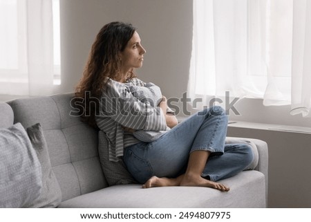 Serious pensive young woman thinking on problem, hugs cushion seated on couch at home, deep on sad thoughts, staring out window, looks depressed, frustrated, misses her beloved, feels lonely. Solitude