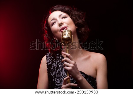 Beautiful young female vocalist in shiny black evening dress singing holding golden vintage microphone, during live musical show
