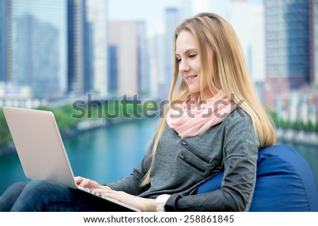 Young girl using laptop, study, working, communicating, ordering online, sitting in comfortable beanbag chair, cityscape on the background
