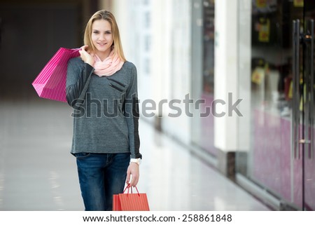 Smiling beautiful teenage girl walking in shopping center with colorful paper bags, passing shopwindow with discount offers, copyspace