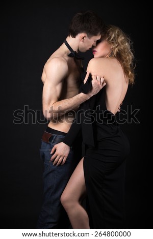 Steamy scene, beautiful sexy young couple foreplay, half naked man in jeans and bow tie taking off his lady evening dress after posh party, studio, black background