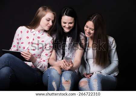 Group of beautiful girlfriends sitting with different devices, looking together at one of the mobile phones and laughing, best friends having fun