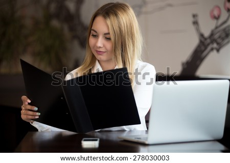 Young beautiful caucasian office woman in formal trendy wear holding case, looking at documents at workplace or menu at cafe, working on laptop