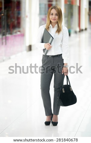 Full length portrait of office employee, beautiful young woman in white shirt holding black document folder in contemporary large building