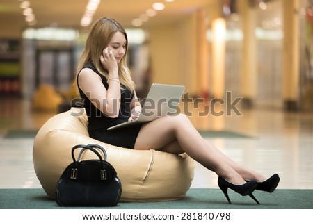 Young serious female sitting on bean bag working on laptop in public wifi area, typing, wearing short black dress, high heel shoes, making call, talking on cellphone in shopping center