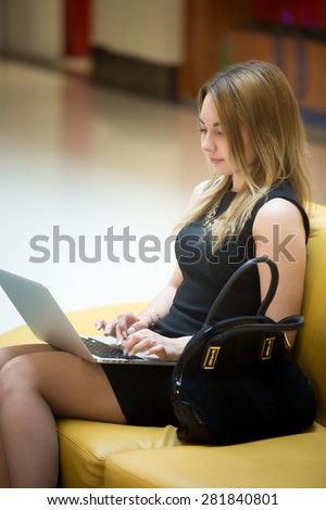Young beautiful female sitting on yellow coach working on laptop in public wifi area, typing