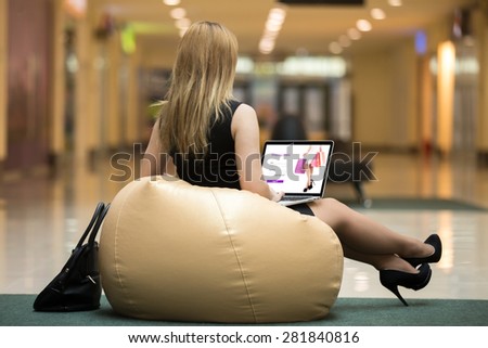Young female wearing short black dress, high heel shoes sitting on bean bag using laptop in public wifi area, ordering online with electronic app, signing in on website, rear view