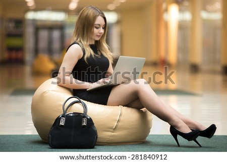Young serious female sitting on beanbag working on laptop in public wifi area, typing