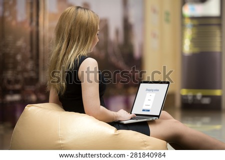 Young attractive female sitting on beanbag using laptop in public wifi area, shopping online, using electronic application, signing in on website