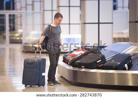 Young handsome man passenger in his 20s waiting to pick luggage at baggage conveyor in arrivals lounge of airport terminal building