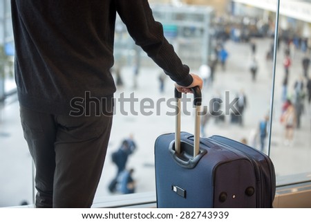 Young handsome man in 20s waiting for flight, standing in modern airport terminal with crowd on background, holding handle of luggage bag, wearing casual clothes, rear view, close up